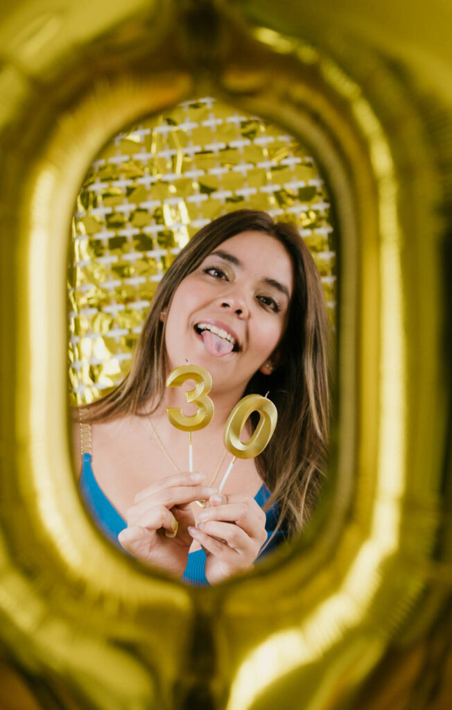 Woman framed in a gold balloon, holding gold "30" candles, playfully sticking out her tongue