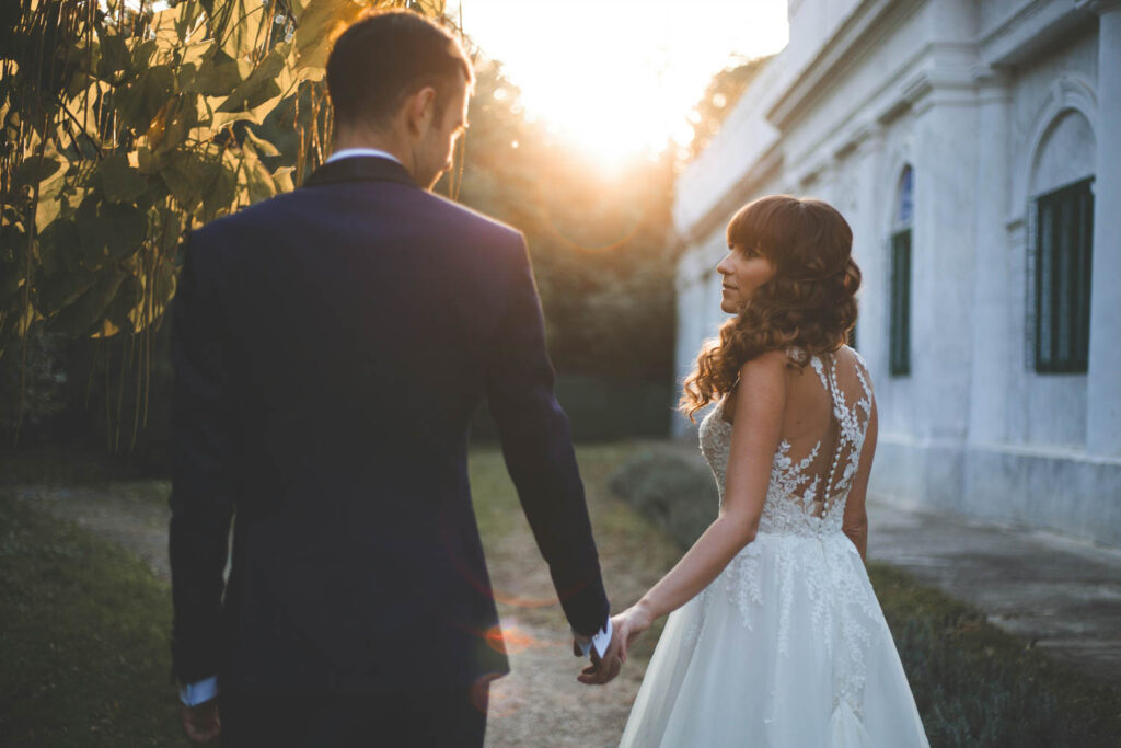 Bride and groom holding hands, walking toward a sunset near a historic building