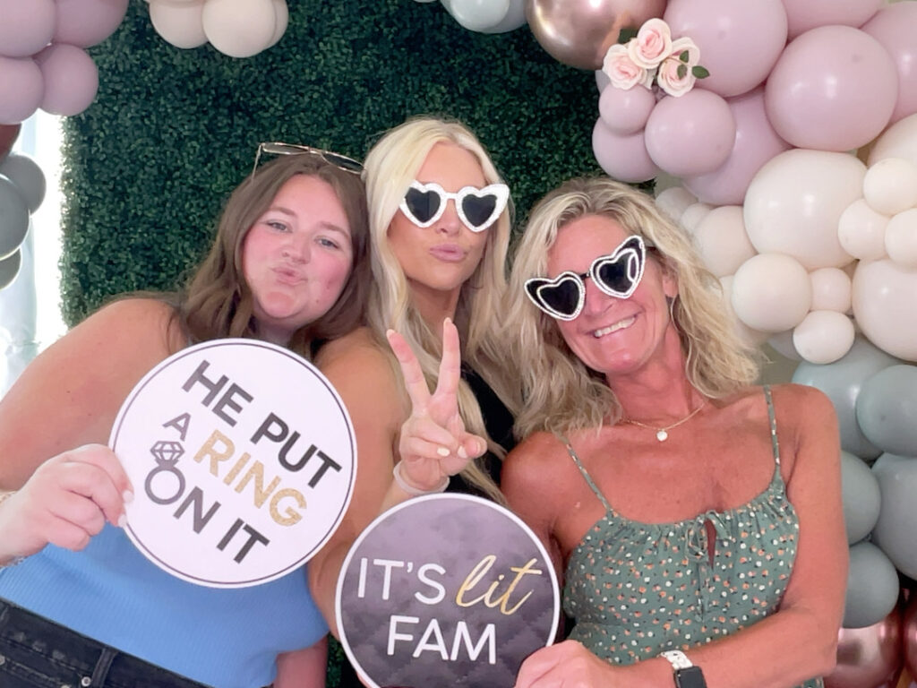 Three women posing with fun props, including heart-shaped glasses and signs