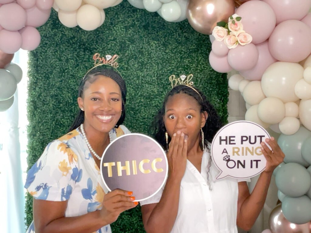 Two women smiling with bride-themed props in front of a balloon-filled backdrop